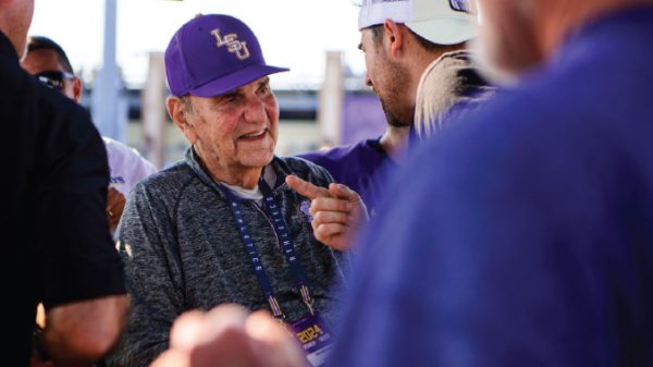 Elderly man in a purple LSU cap smiles while chatting with a younger man at an outdoor gathering.