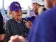 Elderly man in a purple LSU cap smiles while chatting with a younger man at an outdoor gathering.