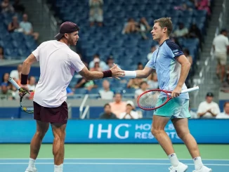 Two male tennis players shake hands at the net after a match on a blue indoor court with spectators in the stands nearby.