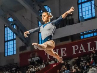 Female gymnast in a blue and white leotard leaps midair during a competition in a gymnasium, with spectators and a red banner in the background.