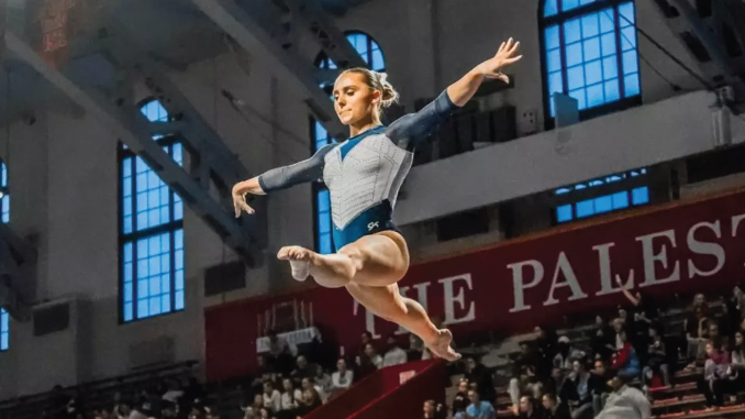 Female gymnast in a blue and white leotard leaps midair during a competition in a gymnasium, with spectators and a red banner in the background.