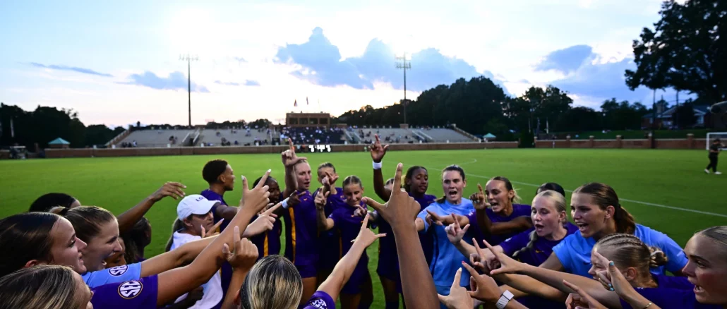 Group of female soccer players in purple uniforms form a huddle with hands raised in the center of a field, stadium stands in the background at dusk.