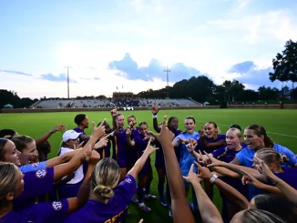 Girls' soccer team in purple uniforms form a circle on the field, hands raised in celebration as crowd watches from the stands behind them at dusk.
