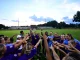 Girls' soccer team in purple uniforms form a circle on the field, hands raised in celebration as crowd watches from the stands behind them at dusk.