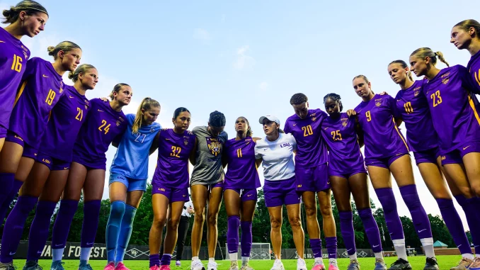 LSU women's soccer players huddling in a circle on the field in purple uniforms.