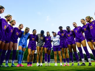 Women’s soccer players in LSU purple uniforms form a circle with arms around each other in a huddle on a sunny field.