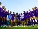 Women’s soccer players in LSU purple uniforms form a circle with arms around each other in a huddle on a sunny field.
