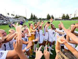 Soccer players in white uniforms celebrate together with raised arms on a sunny field, spectators in the stands behind them.