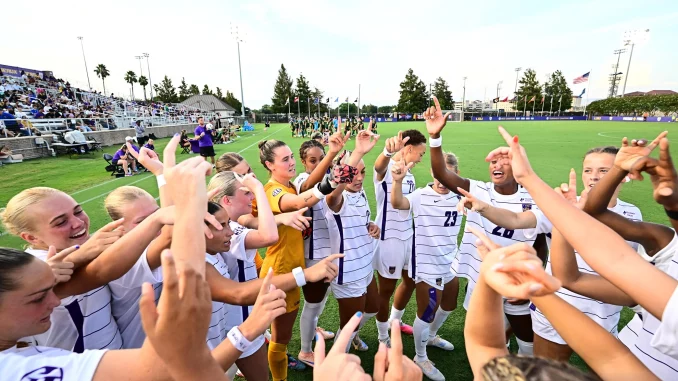 Soccer players in white uniforms celebrate together with raised arms on a sunny field, spectators in the stands behind them.