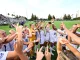 Soccer players in white uniforms celebrate together with raised arms on a sunny field, spectators in the stands behind them.