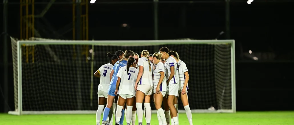 Women’s soccer team huddled in a circle on a well-lit field near a goal at night.
