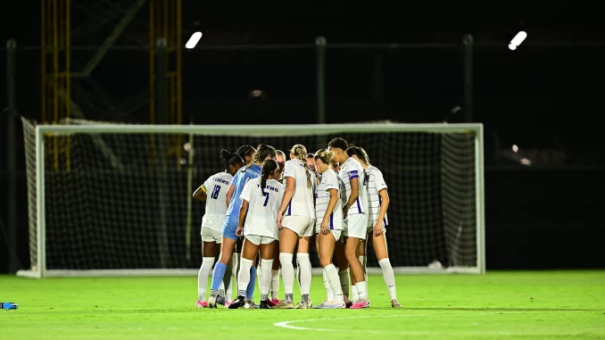 Women’s soccer team huddled in a circle on a well-lit field near a goal at night.