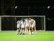 Women’s soccer team huddled in a circle on a well-lit field near a goal at night.