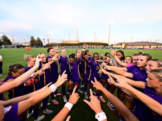 Soccer players in purple uniforms form a circle on the field, hands joined in a team huddle before the game.