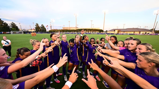 Soccer players in purple uniforms form a circle on the field, hands joined in a team huddle before the game.