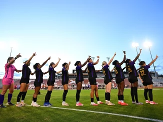 Line of female soccer players in purple and black uniforms stand in a row on the field, each with an arm raised and pointing upward toward the sky at a stadium.