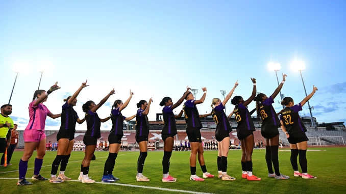 Line of female soccer players in purple and black uniforms stand in a row on the field, each with an arm raised and pointing upward toward the sky at a stadium.