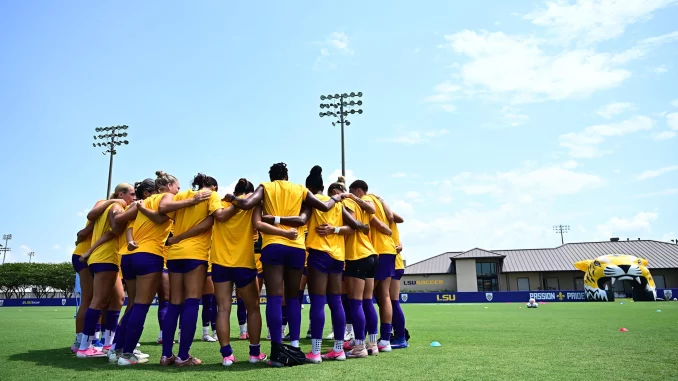 Soccer players in yellow jerseys form a team huddle on a green field under a blue sky at LSU Soccer stadium.