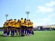 Soccer players in yellow jerseys form a team huddle on a green field under a blue sky at LSU Soccer stadium.