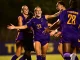 Female soccer players in purple uniforms celebrate together on a night game field, arms raised in joy.