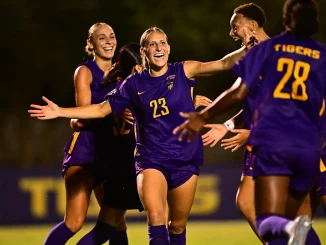 Soccer players in purple uniforms celebrate together on a field at night, arms outstretched in joy.