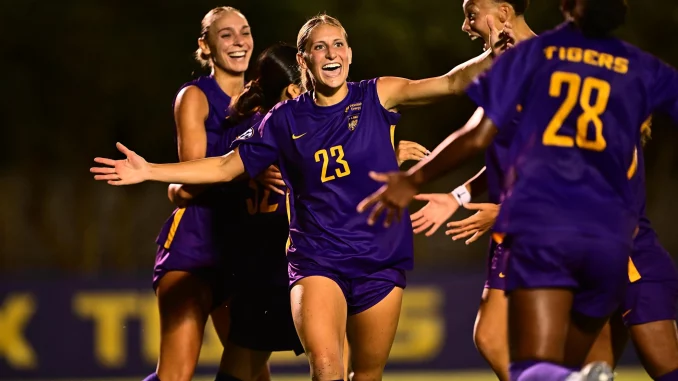 Soccer players in purple uniforms celebrate together on a field at night, arms outstretched in joy.