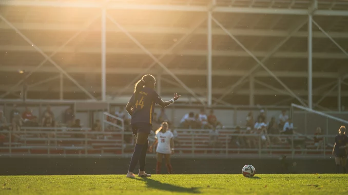 Soccer player with number 14 in a dark uniform on a sunlit field, ball at her feet, stands and spectators in the background.