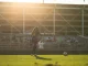 Soccer player with number 14 in a dark uniform on a sunlit field, ball at her feet, stands and spectators in the background.