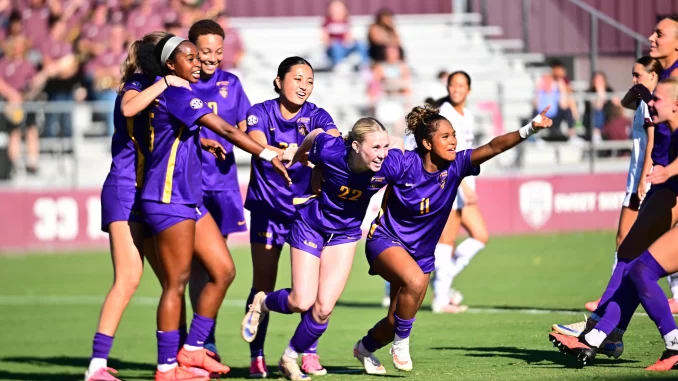 Soccer players in purple uniforms celebrate on the field, arms linked and smiling as a group hug.