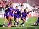 Soccer players in purple uniforms celebrate on the field, arms linked and smiling as a group hug.