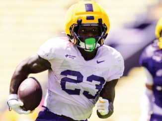 Football player wearing a yellow helmet and white jersey with number 23 runs with the ball on a sunny field, opponent in purple behind him