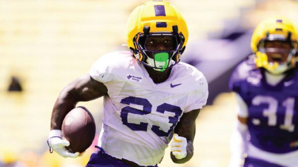 Football player wearing a yellow helmet and white jersey with number 23 runs with the ball on a sunny field, opponent in purple behind him