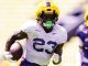 Football player wearing a yellow helmet and white jersey with number 23 runs with the ball on a sunny field, opponent in purple behind him