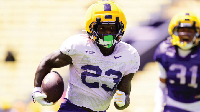 Football player wearing a yellow helmet and white jersey with number 23 runs with the ball on a sunny field, opponent in purple behind him