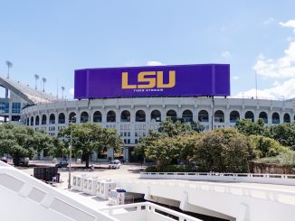 Exterior view of LSU's Tiger Stadium with a large purple LSU sign on top against a blue sky.