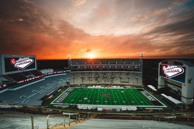 Mississippi State football stadium at sunset, empty stands with a green field and illuminated video boards reading 'Mississippi State Starkvegas'.