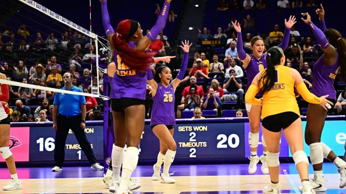 LSU women's volleyball players in purple celebrate a point on the court as fans cheer in the stands behind them.