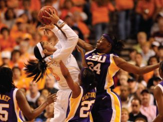 Women’s college basketball action as a white-uniform player shoots while purple-clad defenders crowd her under a packed crowd wearing orange.