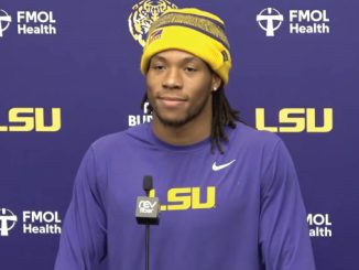 Athlete in a purple LSU shirt and yellow beanie speaking at a press conference, microphone in front, backdrop with LSU logos.