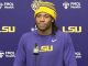 Athlete in a purple LSU shirt and yellow beanie speaking at a press conference, microphone in front, backdrop with LSU logos.