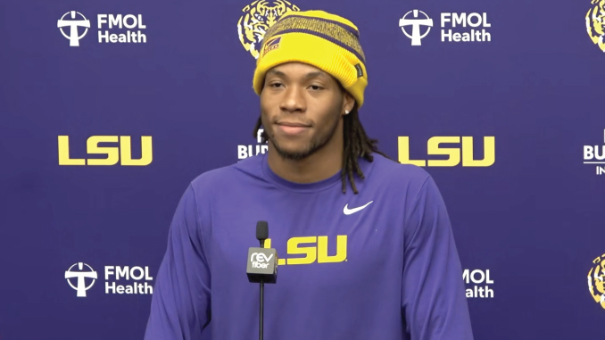 Athlete in a purple LSU shirt and yellow beanie speaking at a press conference, microphone in front, backdrop with LSU logos.