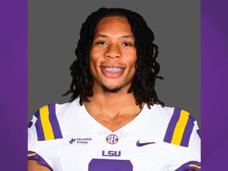 Portrait of an LSU football player in a white jersey with purple and gold stripes, smiling with braces against a gray backdrop.