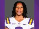 Portrait of an LSU football player in a white jersey with purple and gold stripes, smiling with braces against a gray backdrop.