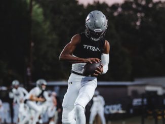 Football player in white uniform runs with ball tucked under arm during a game, helmet on, teammates blurred in the background.