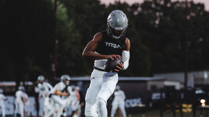 Football player in white uniform runs with ball tucked under arm during a game, helmet on, teammates blurred in the background.