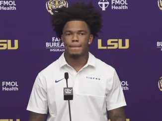 Young man with voluminous hair in a white polo at a podium during an LSU press conference against a purple backdrop with LSU and sponsor logos