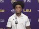 Young man with voluminous hair in a white polo at a podium during an LSU press conference against a purple backdrop with LSU and sponsor logos