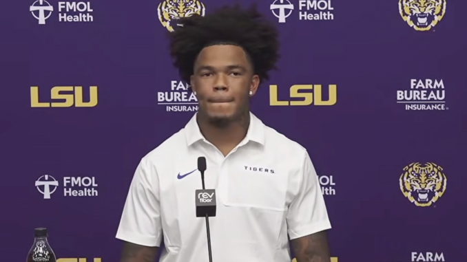 Young man with voluminous hair in a white polo at a podium during an LSU press conference against a purple backdrop with LSU and sponsor logos