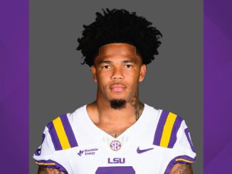 Portrait of an LSU football player in a white jersey with purple and gold stripes, facing the camera against a gray backdrop.