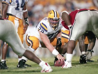 LSU offensive lineman snaps the ball at the line of scrimmage during a football play, surrounded by teammates in white and purple uniforms